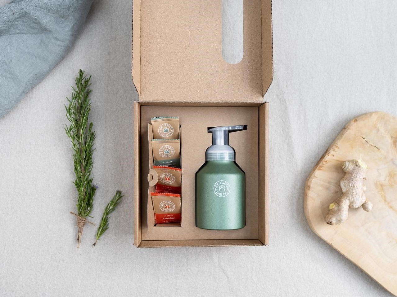 The Green Lab Co refill kit with a reusable aluminum pump bottle and tablet sachets in a cardboard box, styled with rosemary and ginger.